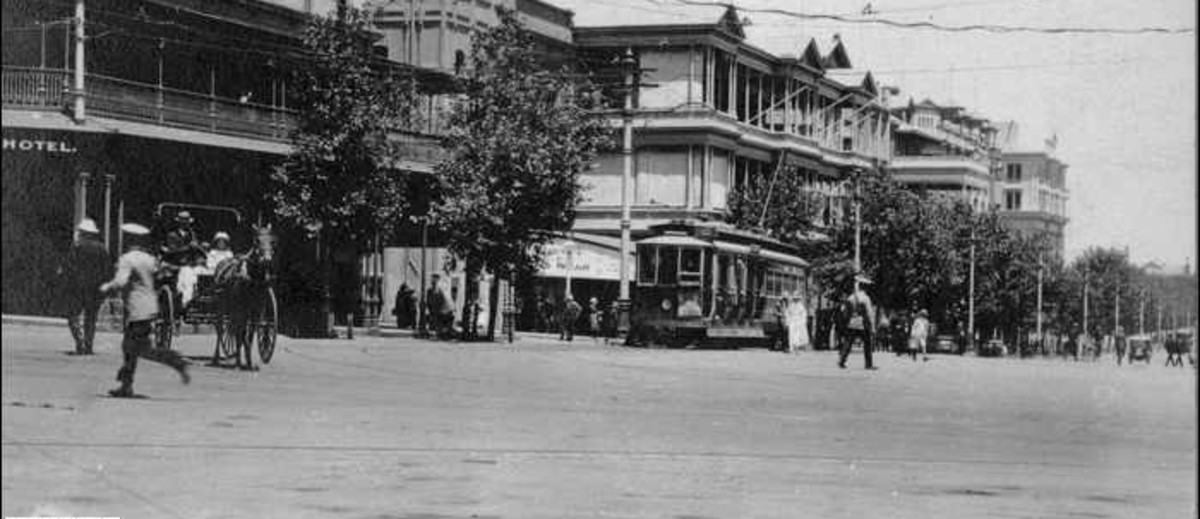 Image: a series of three storey buildings line one side of a wide city street. Between the buildings and the road is a line of trees. Travelling along the street are horse-drawn vehicles, motor cars and an electric tram, as well as people in 1920s clothes