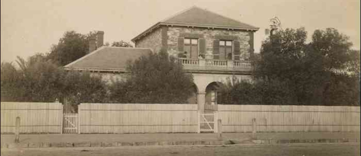 Image: a two storey stone house behind a high fence and partly obscured by trees. The second story windows are shuttered and an arched verandah supports a balcony with stone balustrade.