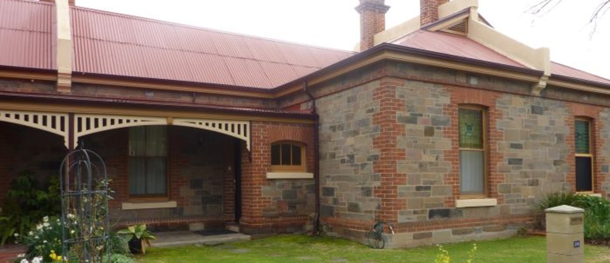 Image: A single storey bluestone building with red brick quoins, red tin roof with cream details and a number of brick chimneys. The building also has a verandah and decorative stained glass on two of its windows. 