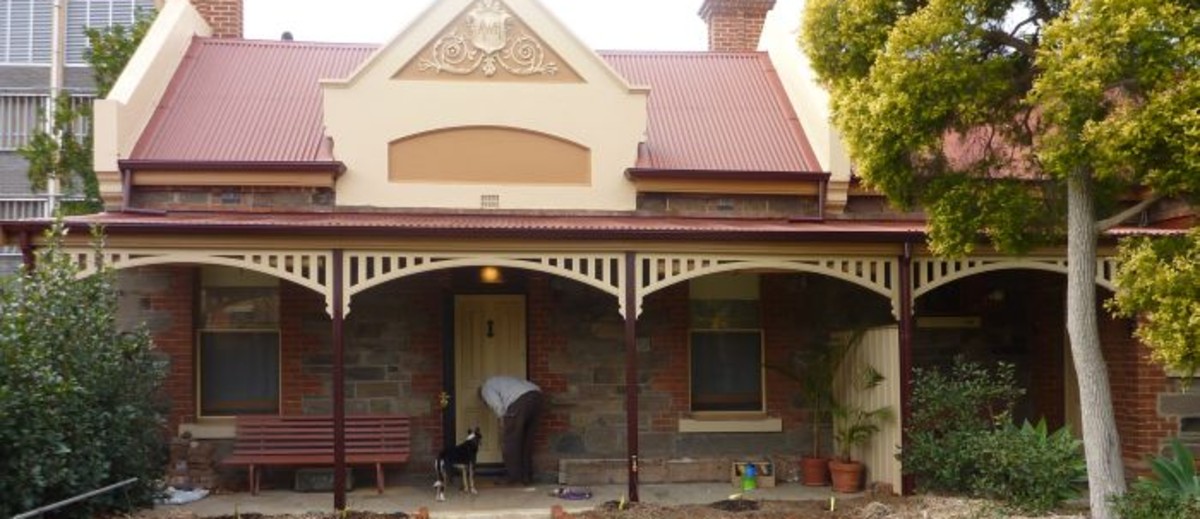 Image: A man and his dog stand outside a bluestone building with red brick quoins around its windows and two red brick chimneys. The building also has a red tin roof, simple wooden verandah and a gable with decorative moulding painted in cream and taupe. 