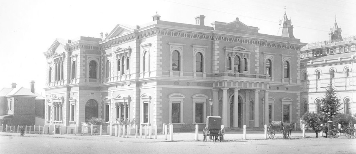 Image: A large, two-storey building fronted by a dirt street and two horse-drawn carts