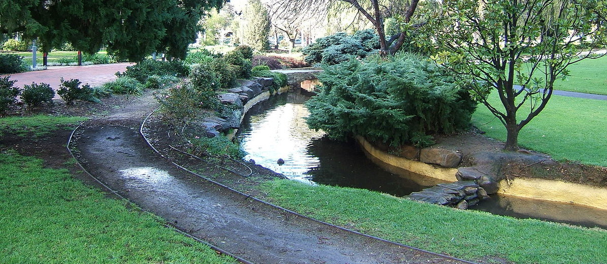 Image: A creek winds through a landscaped wooded parkland. A footpath follows the course of the creek