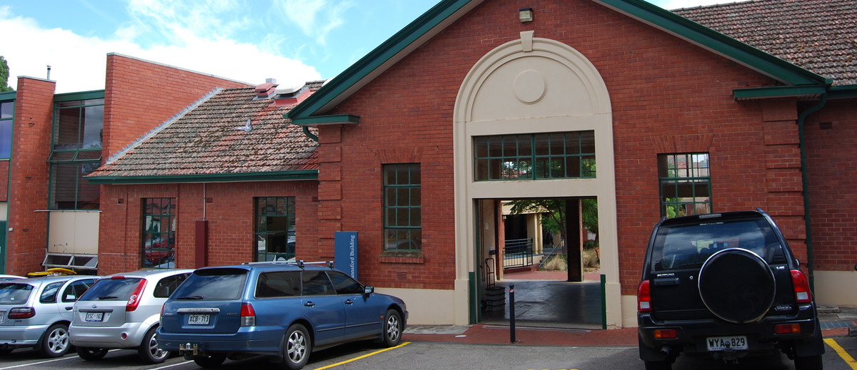 Image: The front of a single-storey brick building with a white arch entranceway. Several modern auto-mobiles are parked in front of the building