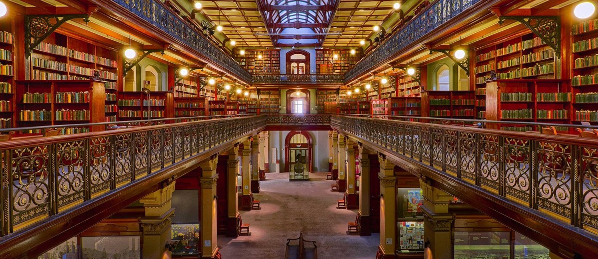 Image: The interior of a large, three-storey Victorian-era library. A large skylight in the roof illuminates the building’s three floors of bookshelves and museum exhibits
