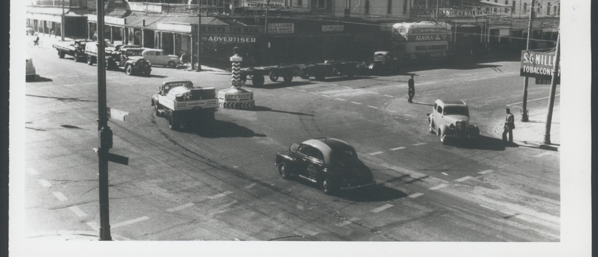 Image: Various mid-twentieth century auto-mobiles pass through an intersection in an urban area. A large plinth stands in the centre of the intersection and is marked with the words ‘Keep Left’ and left-facing arrows 