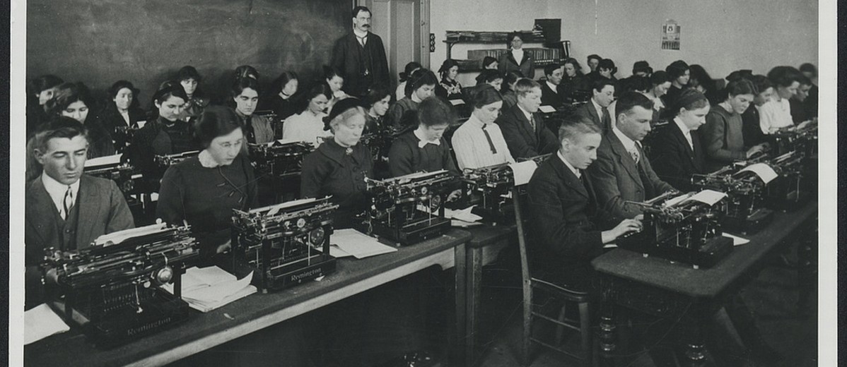 Image: A group of Caucasian men and women dressed in early twentieth century attire practice typing in a classroom. A moustachioed male Caucasian instructor looks on in the background