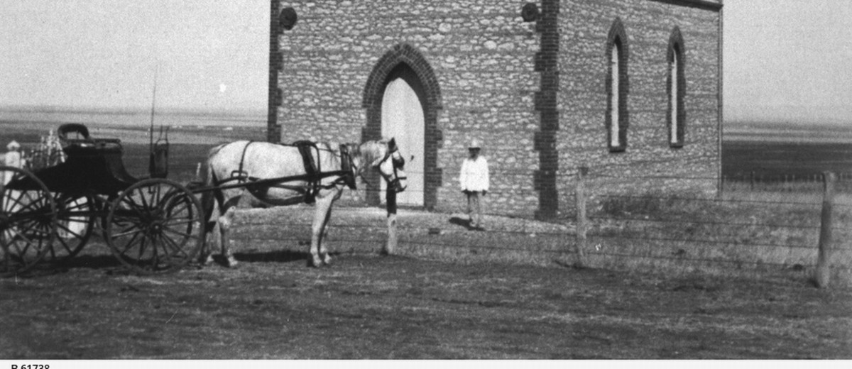 Image: A Caucasian man in a hat and light-coloured top stands outside the front door of a small Bluestone chapel in a remote setting. A horse and buggy stand nearby