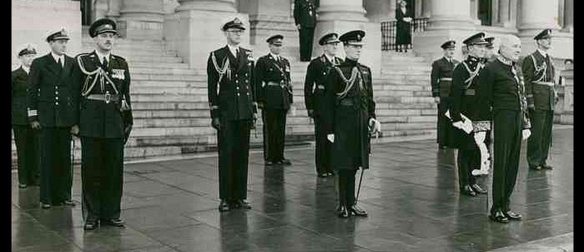 Image: A middle-aged man attired in a ceremonial dress uniform stands at the head of a group of military officers, all of whom are similarly attired. A large building with columns is visible in the immediate background