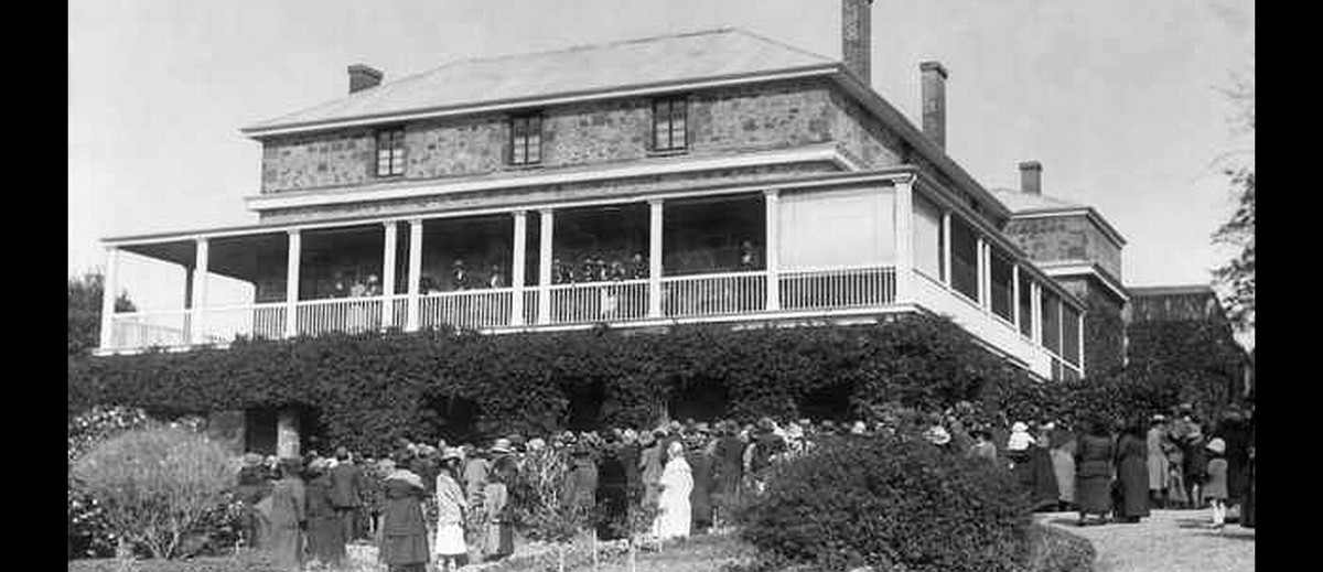 Image: A large, three-storey stone stately home with wraparound second-storey verandah. A group of people are gathered outside at the front of the house