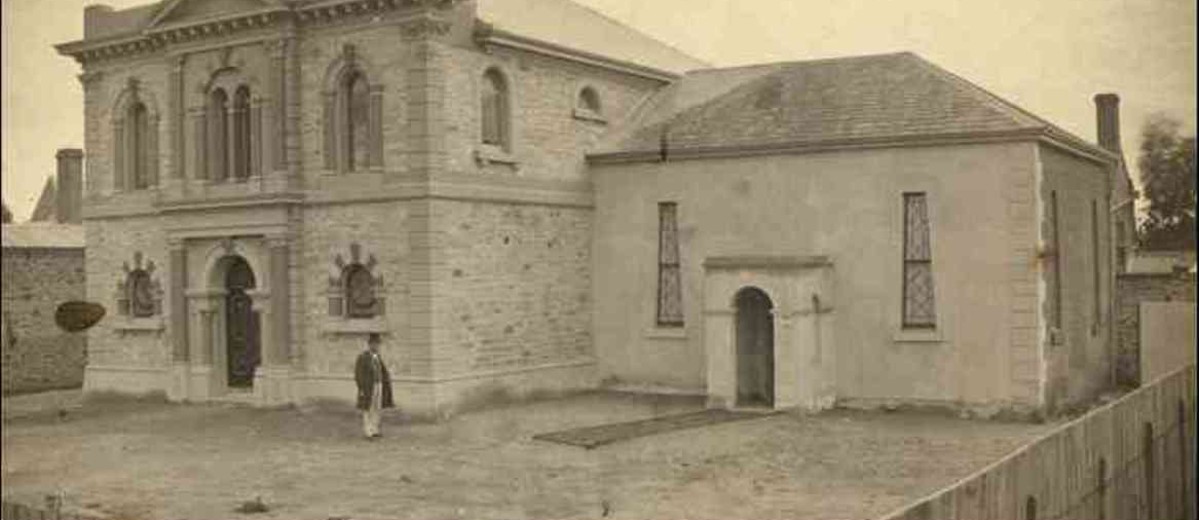 Image: A man in a top-hat and Victorian clothing stands in front of a large, two-storey rectangular stone building