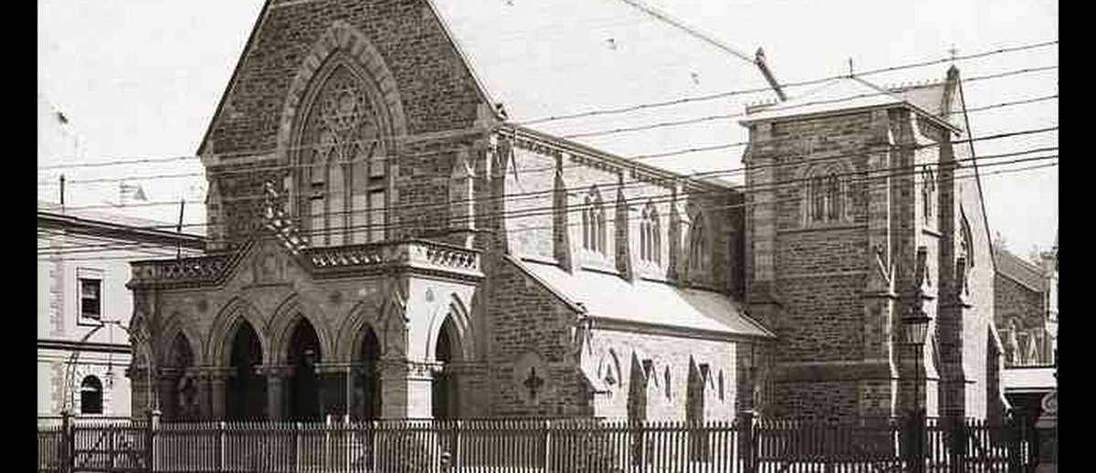 Image: A large bluestone church is surrounded on three sides by a picket fence and borders a road. Electric trolley lines are visible in front of the church