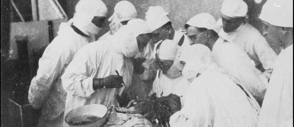 Image: A group of surgeons and medical technicians in white surgery gowns surround a patient on an operating table. Three men closest to the patient are performing an operation