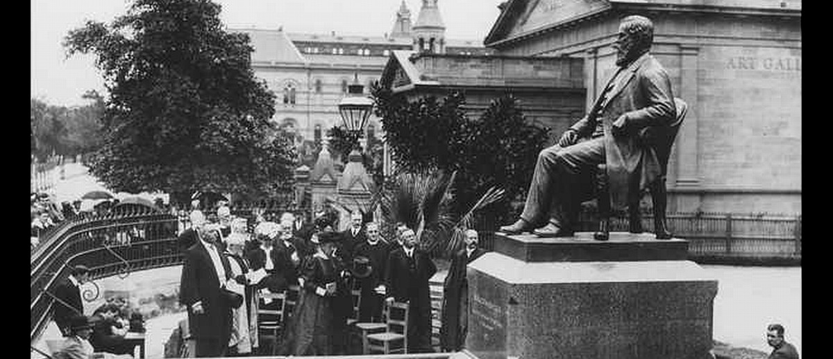 Image: A crowd of people in Edwardian-era attire gather around a large bronze statue of a bearded man sitting in a chair