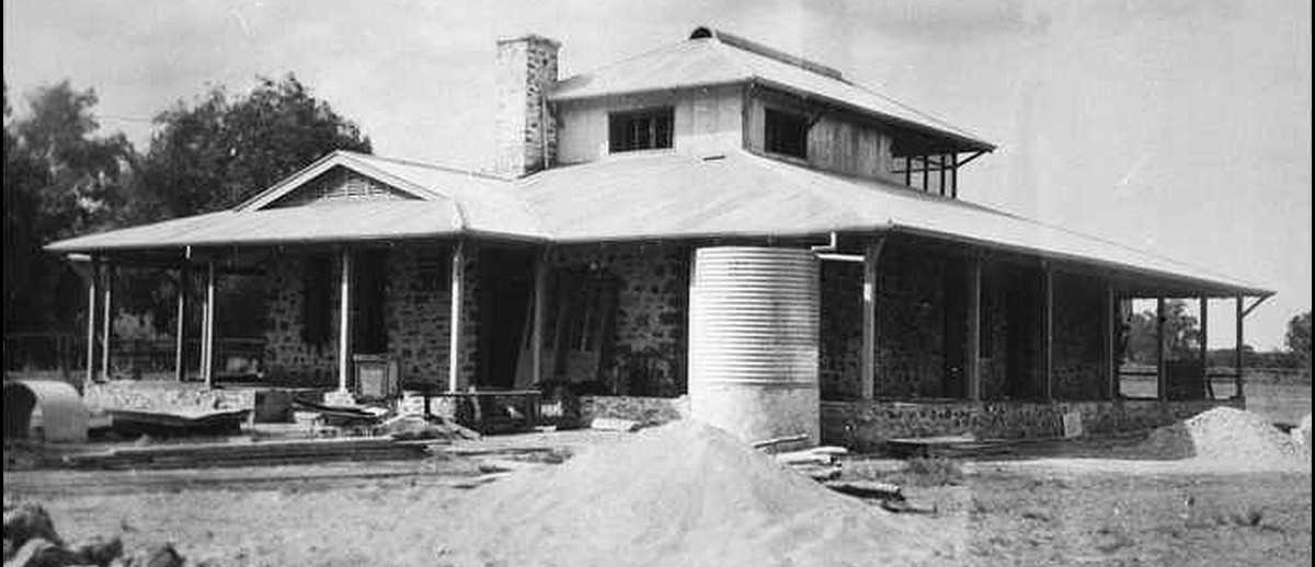 Image: A single-storey stone building with gabled roof and wraparound verandah. A corrugated metal water tank and evidence of construction is visible in the foreground
