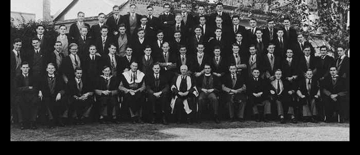 Image: A group of men in mid-20th century suits and academic gowns pose for a photograph outside a large, singles-storey stone building