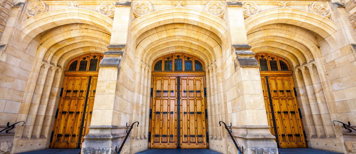 Image: The entrance to an ornate, historic building made of stone. Three large wooden doors are contained within corresponding stone archways