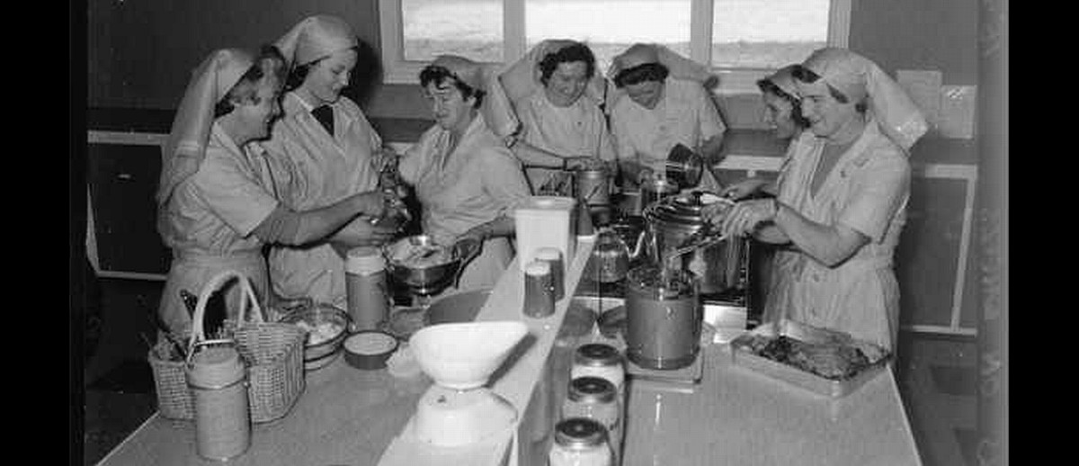Image: A group of seven Caucasian women in 1960s attire that resemble nursing uniforms prepare and pack food in a small kitchen