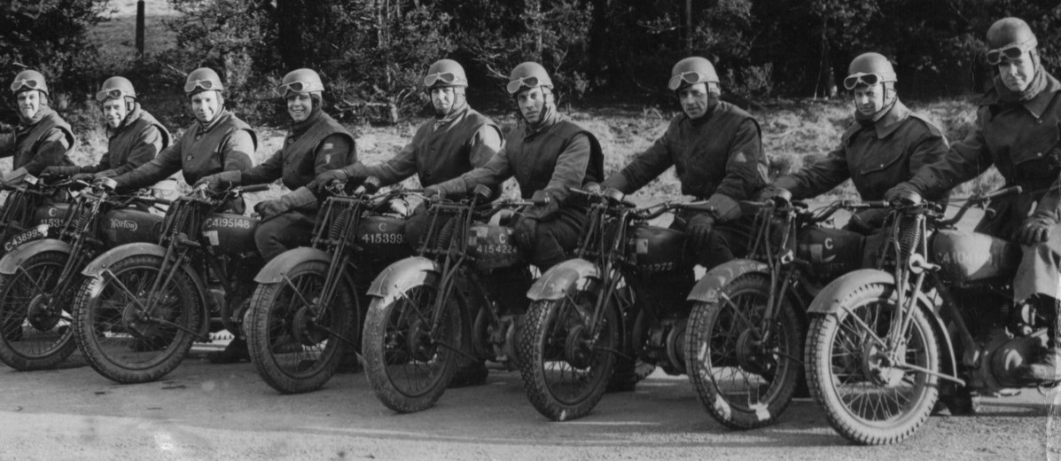 Image: Nine men wearing Second World War-era military uniforms each sits astride a motorcycle. Each man is wearing a helmet with goggles, and all are arranged in a line abreast of one another