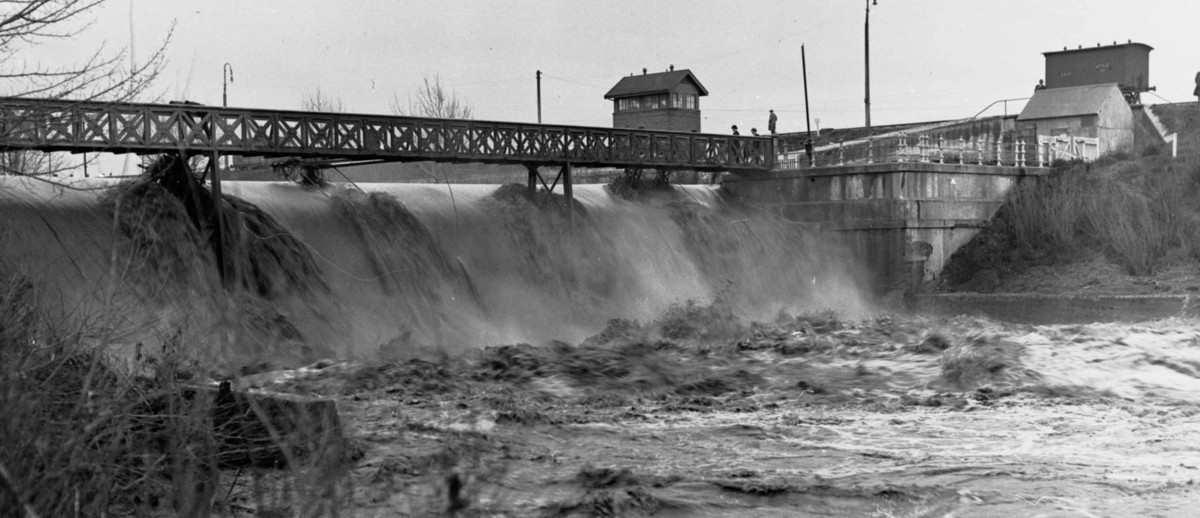 Image: A torrent of flood water pours through a weir along a river. Deep flood waters are visible flowing downstream in the foreground, and a group of men watch the flood from the top of the weir