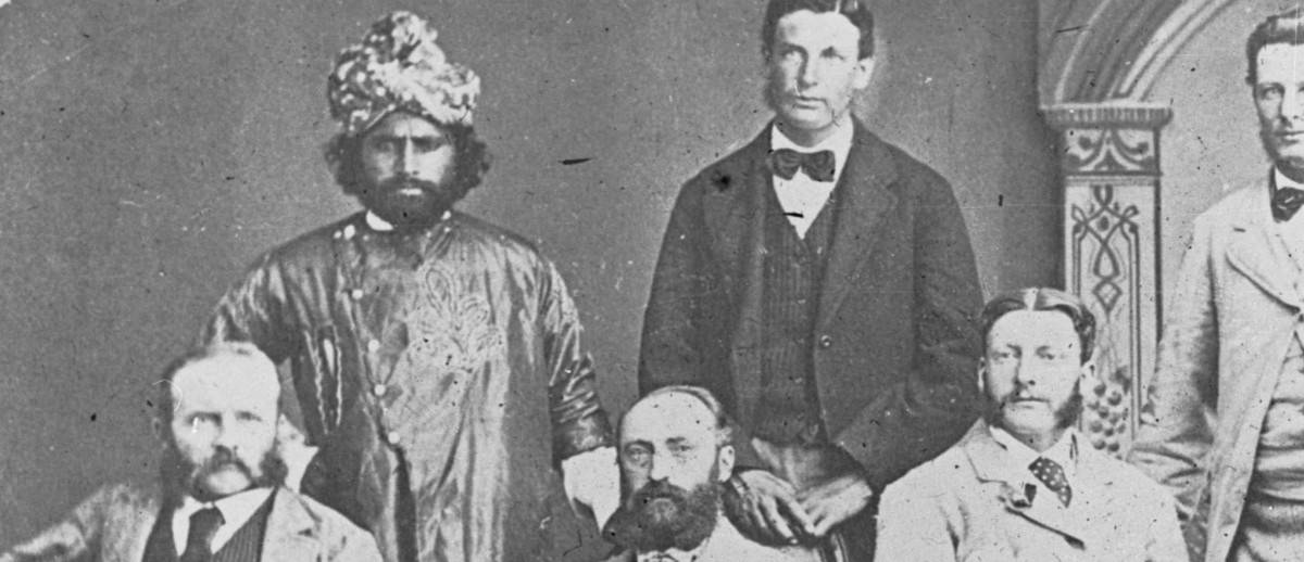 Image: A group of seven men, including an Afghan cameleer and Aboriginal tracker, pose for a photograph. The cameleer is dressed in ornate traditional costume with a turban