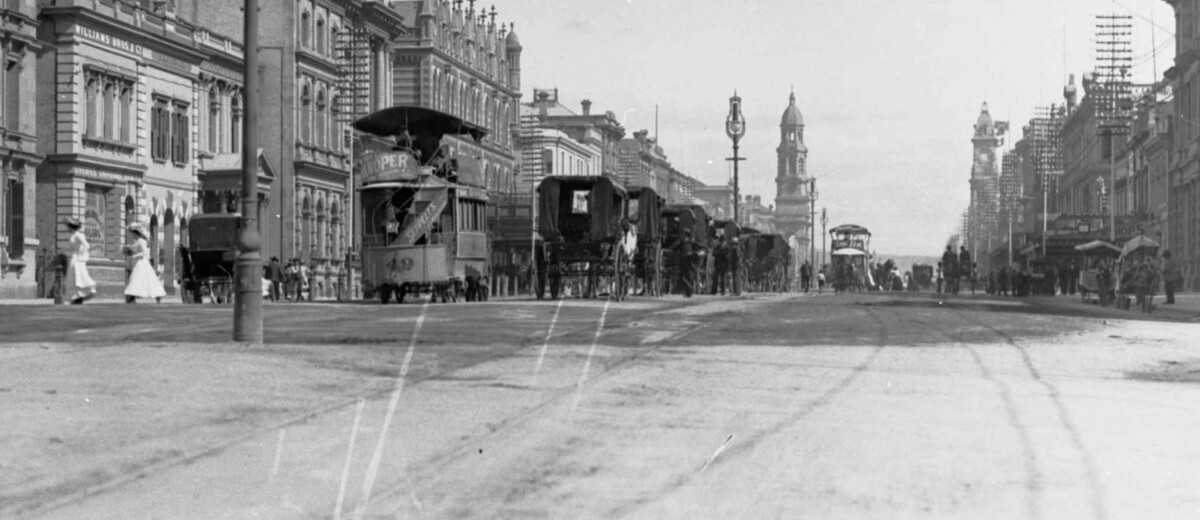 Image: black and white photograph of city street, with tram, horse drawn vehicles and pedestrians