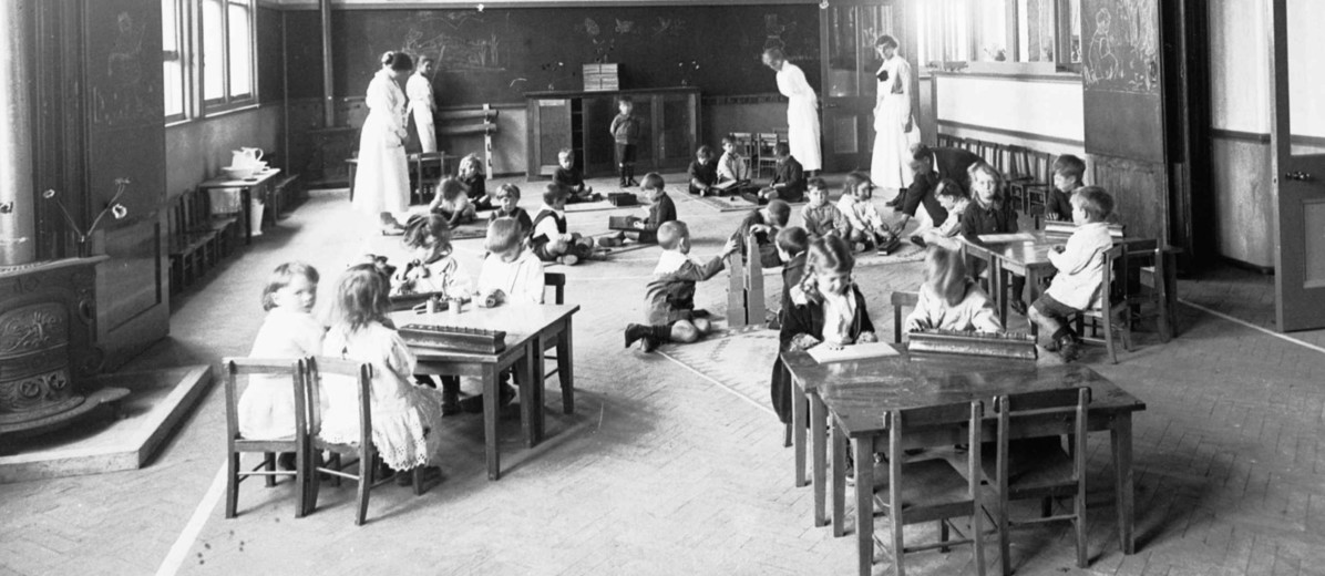 Image: A group of small children play in a large open room while four female teachers in Edwardian attire look on