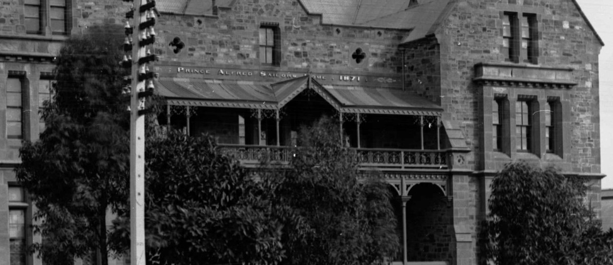 Image: A large, three-storey mid-nineteenth century bluestone building fronted by a dirt street. A sign reading ‘Prince Alfred Sailors’ Home, 1871’ is visible above the front entrance
