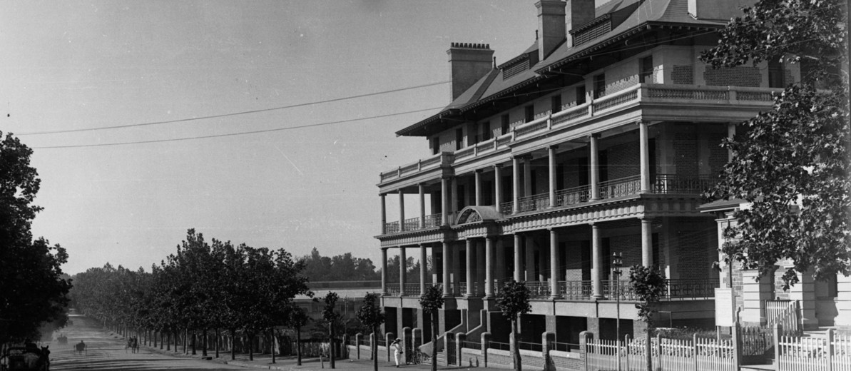 Image: A large, four-storey brick building sits alongside a dirt road lined with trees. Four horse-drawn carts are visible travelling along the street