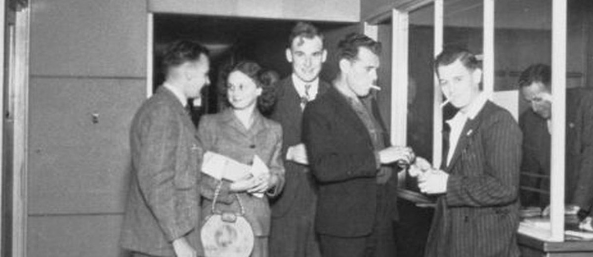 Image: six men and a woman in 1940s era dress stand around a reception desk while a seventh man leans on the desk, writing, behind a glass barrier. In the foreground a second woman in a hat sits looking at the group.
