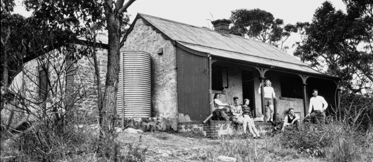 Image: A small group of young men and women sit on the porch of an old stone cottage. The cottage is surrounded by trees and scrub