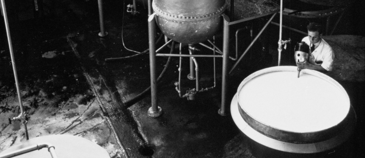Image: A young Caucasian man in a white laboratory coat uses an industrial mixer to combine chemicals in a large steel vat. Large metal storage tanks are visible in the immediate background