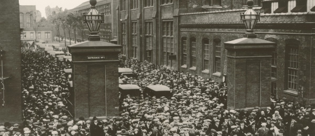 Image: A mass of men and women in 1920s attire walk through the entrance gates of a large, multi-storey brick factory