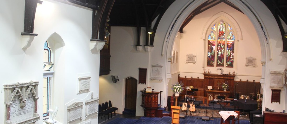 Image: An large, interior room with a high ceiling, wooden pews and a large stained glass window