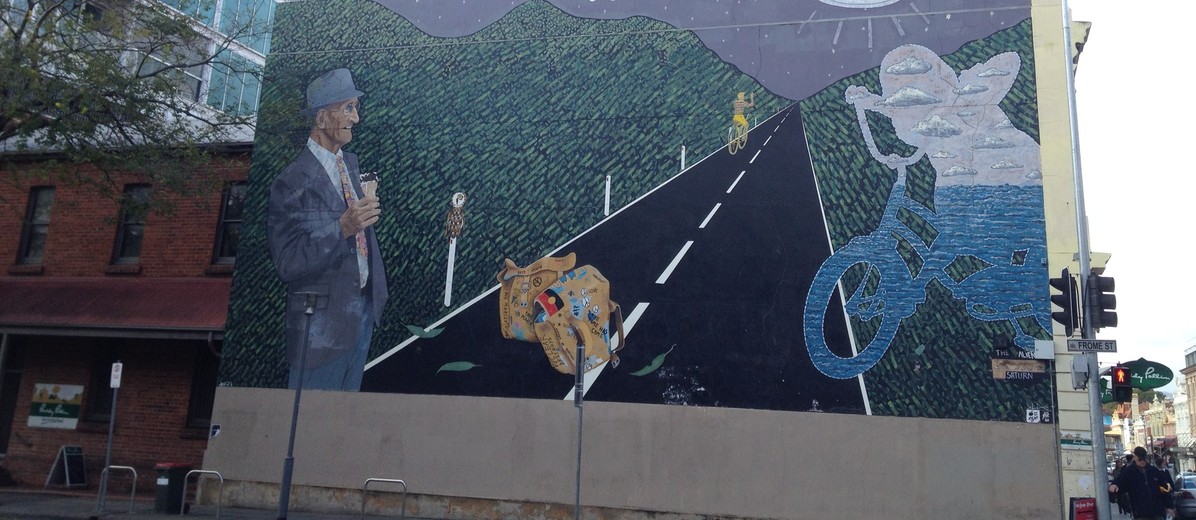 Image: mural of an older man with an ice cream cone watching an abstract figure and a boy both riding bikes on a road in the moonlight. Above, two aliens are on an unidentified flying object