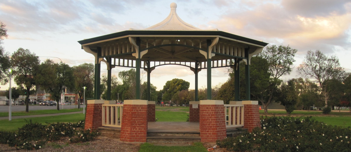 Image: A green and white gazebo with red brick footers stands in a park. A street with cars is visible in the left background