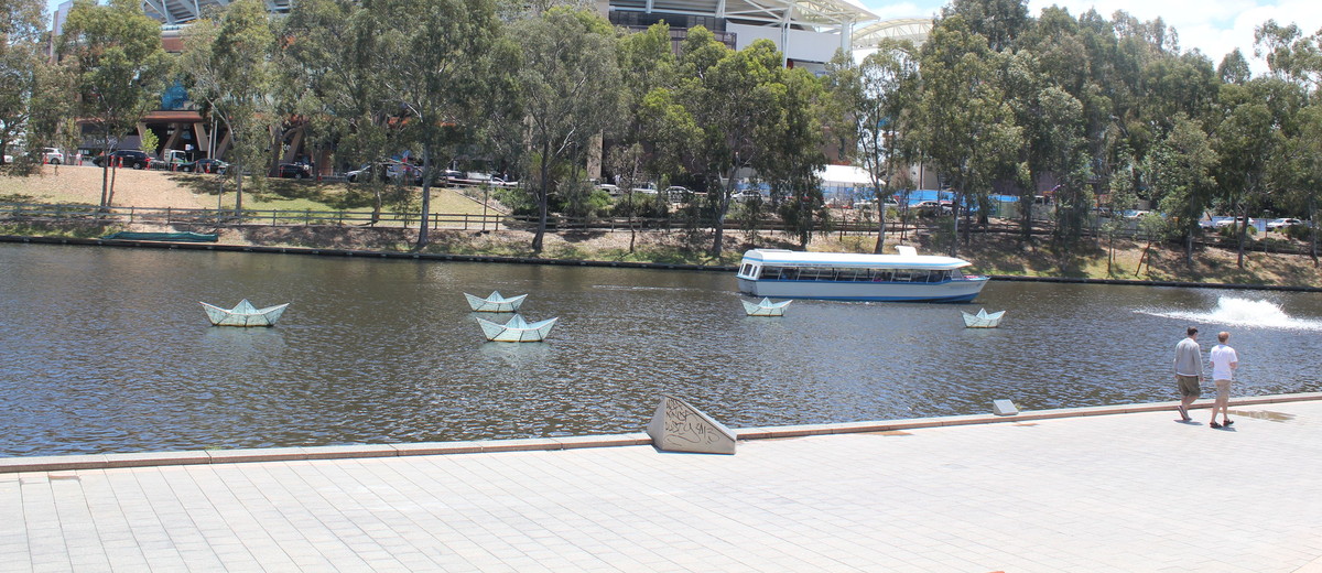 Image: a long, narrow blue and white boat with a roof sails past a stadium.