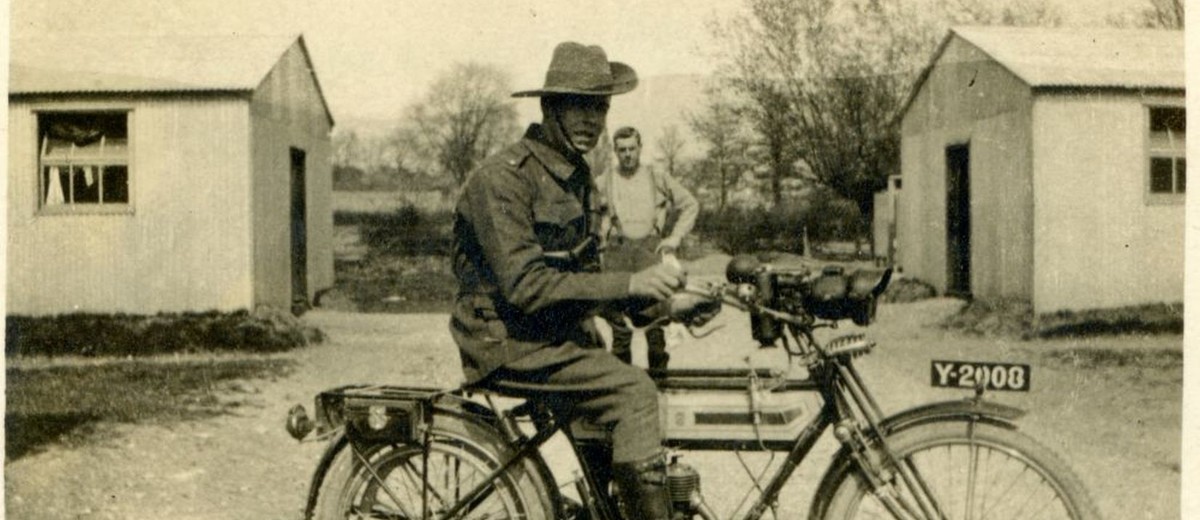 Sepia photo of a man in military uniform on a motorcycle. 