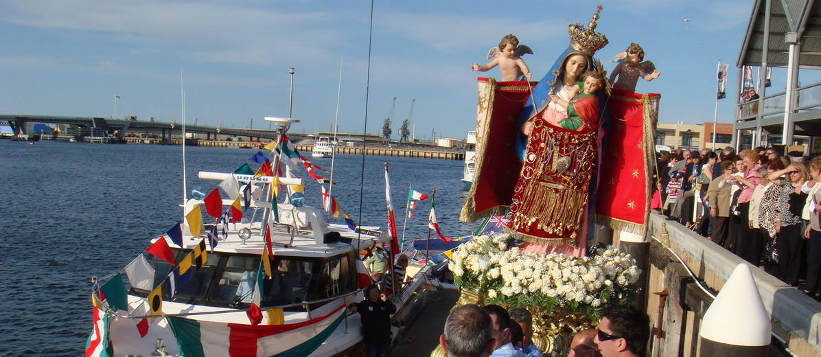 Image: A group of people watch men in Catholic vestments carry an effigy of the Madonna and Child to a motorboat moored at a wharf