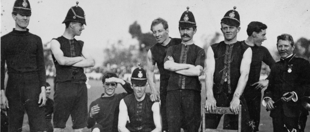 Image: A group of Australian Rules Football players in early twentieth century uniforms pose for a photograph. Most of the men are wearing period police helmets