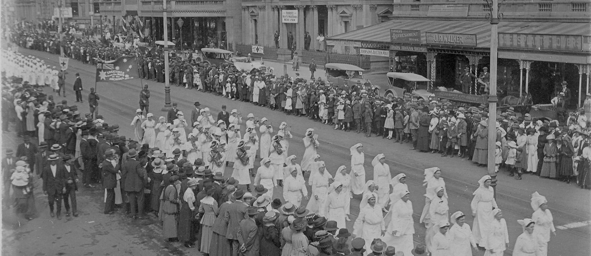 Crowds line street as girls march past, dressed in white with their instruments.