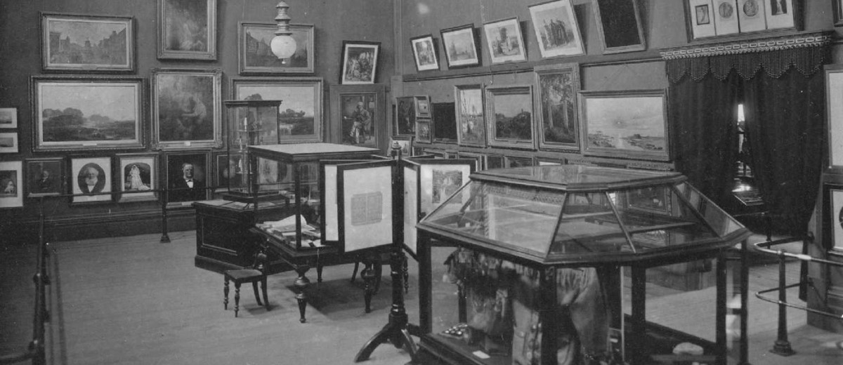 Image: A high-ceilinged room containing several pieces of artwork on its walls and a handful of display cases containing miscellaneous objects
