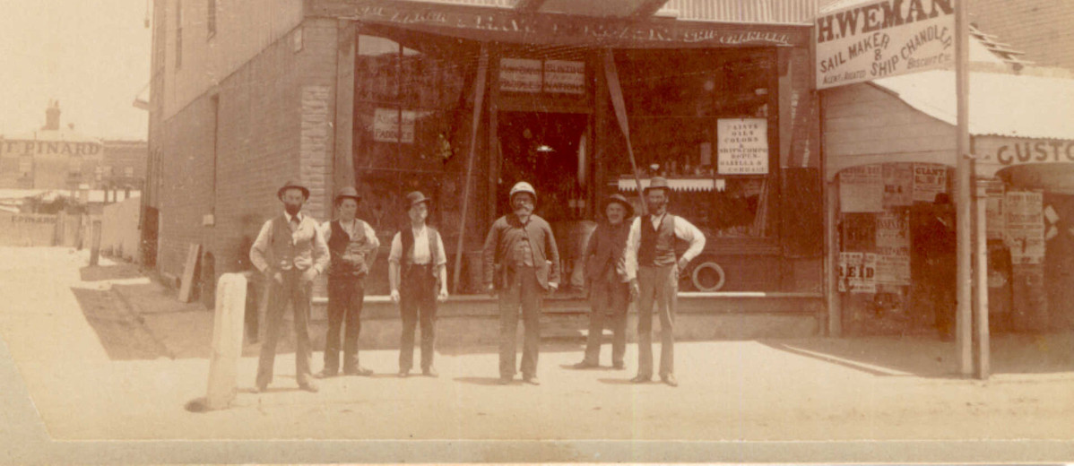 Image: A group of six men of various ages in Victorian attire stand in front of a shop. A painted banner on the top of the shop reads ‘Hy. Weman. Sail Maker & Ship Chandler’