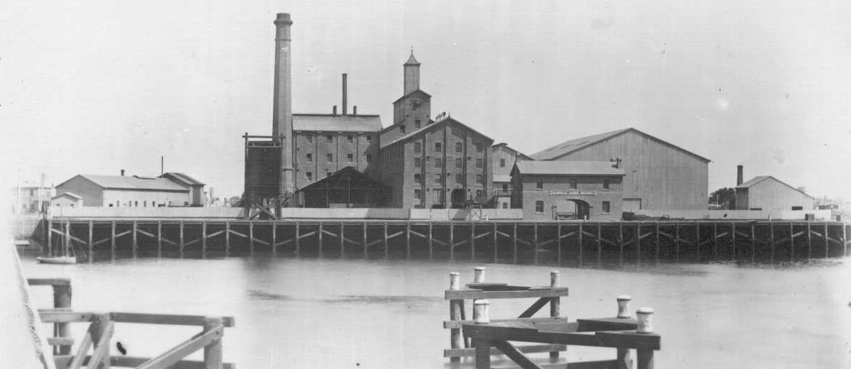 Image: A complex of multi-storey brick buildings and large, corrugated metal-clad warehouses fronted by a river. A large brick chimney is visible at one end of the complex