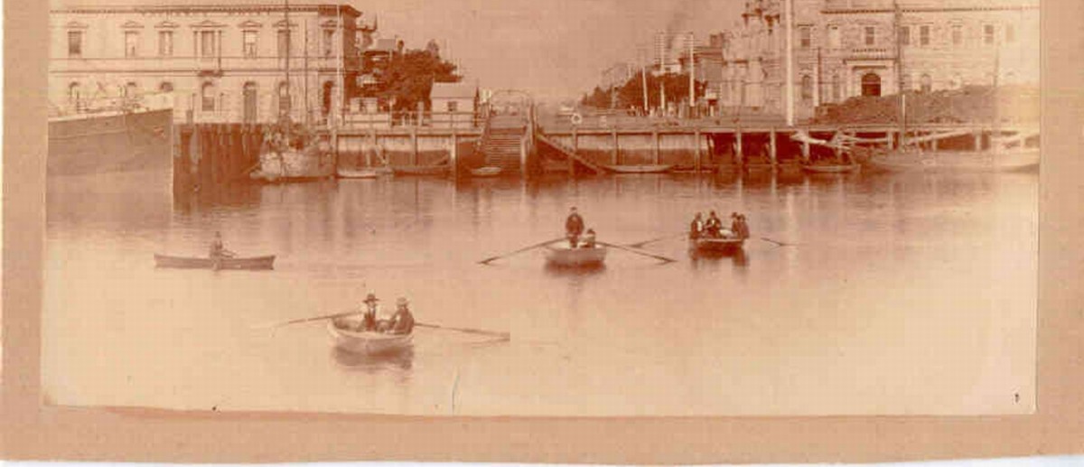 Image: Four rowboats sit unmoving in the middle of a river before a town waterfront. People within the boats are looking at the camera