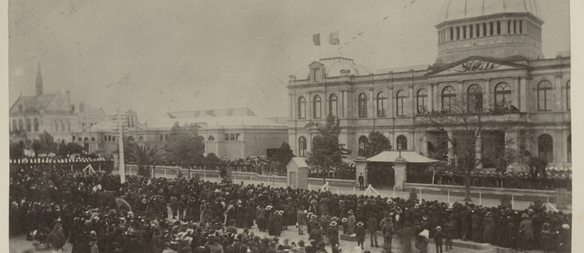 Image: A large group of people in 1880s dress stand in distinct lines in front of a large building with a domed roof which is decorated with flags and bunting. 