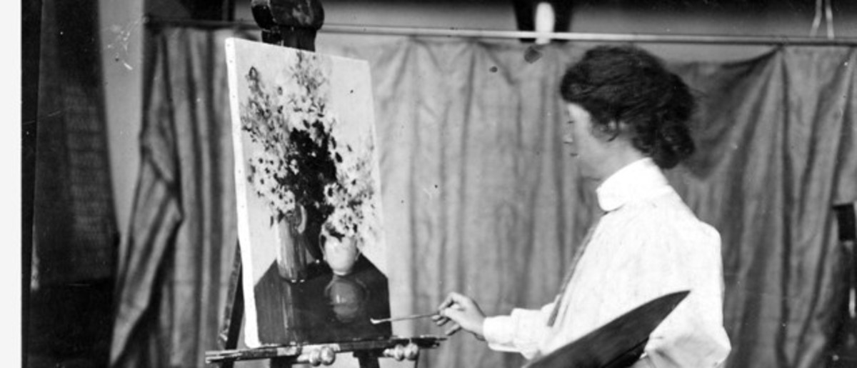 Image: A young Caucasian woman in a full-length Edwardian-era dress paints a still life on an easel. The painting features a couple of vases holding flowers