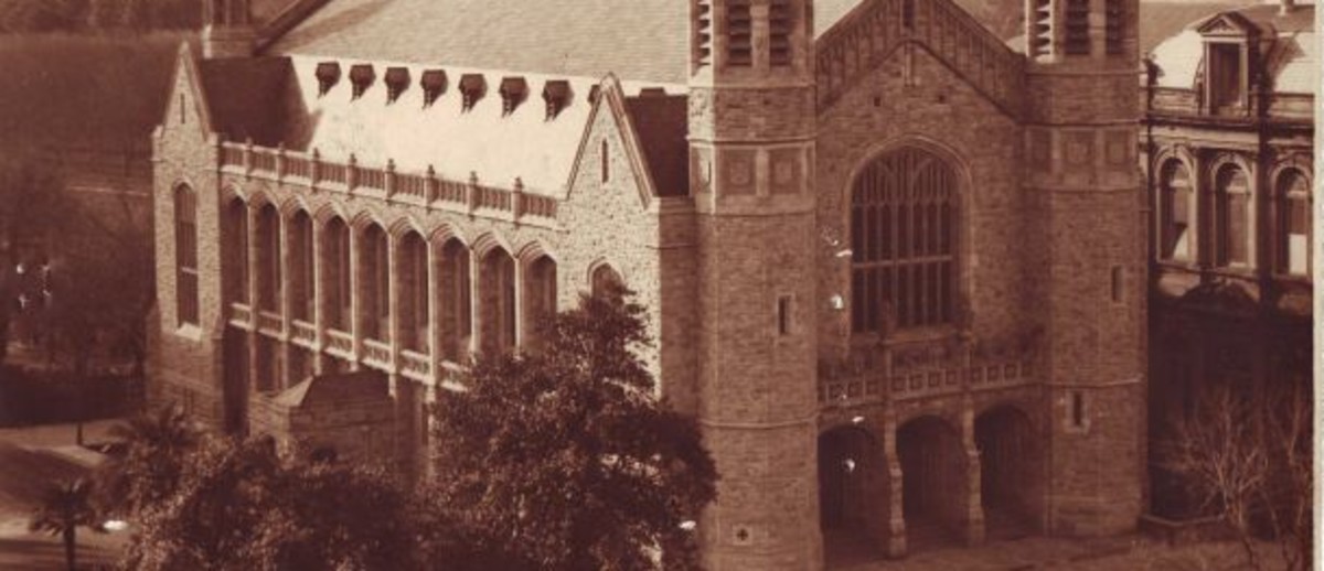 Image: a high angle photograph of a large stone hall with towers and three doors under a large window at the front.