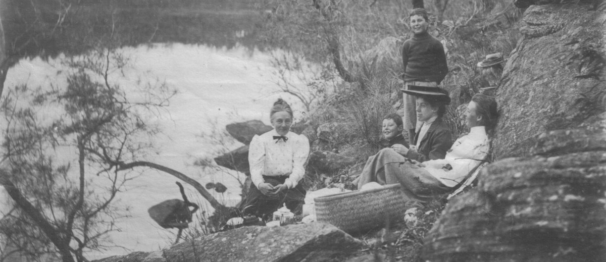 Image: Three women and two boys have a picnic on the banks of a river.