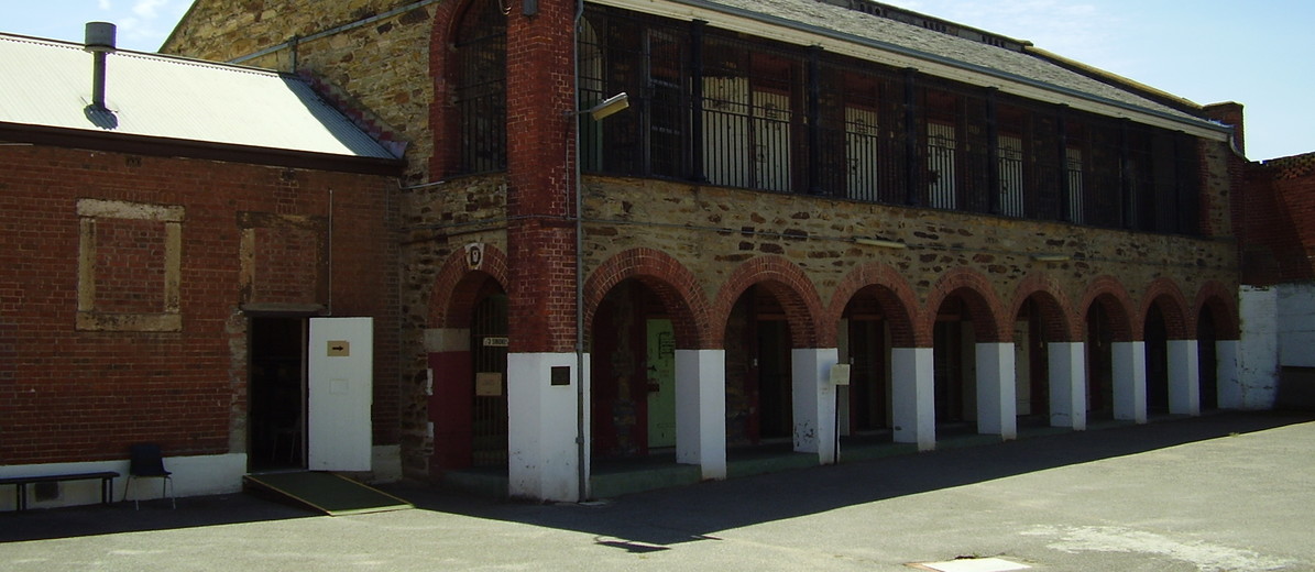 Image: a two storey stone building with a brick arched loggia and barred upper storey balcony. A security camera is mounted on one end of the building.