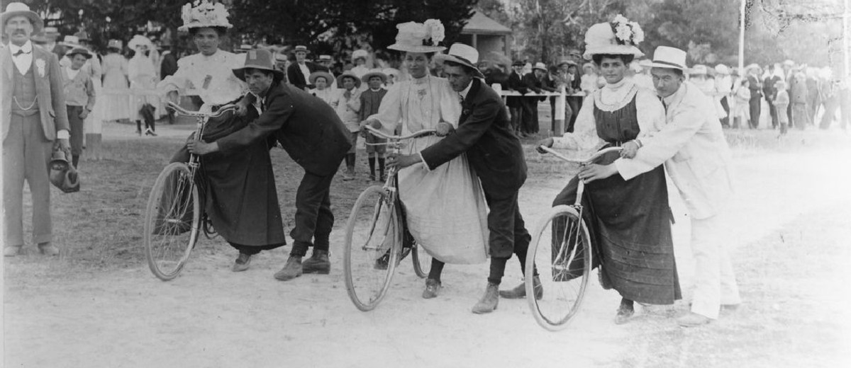 Image: three women on bicycles about to begin a race. Men hold the bicycles steady.